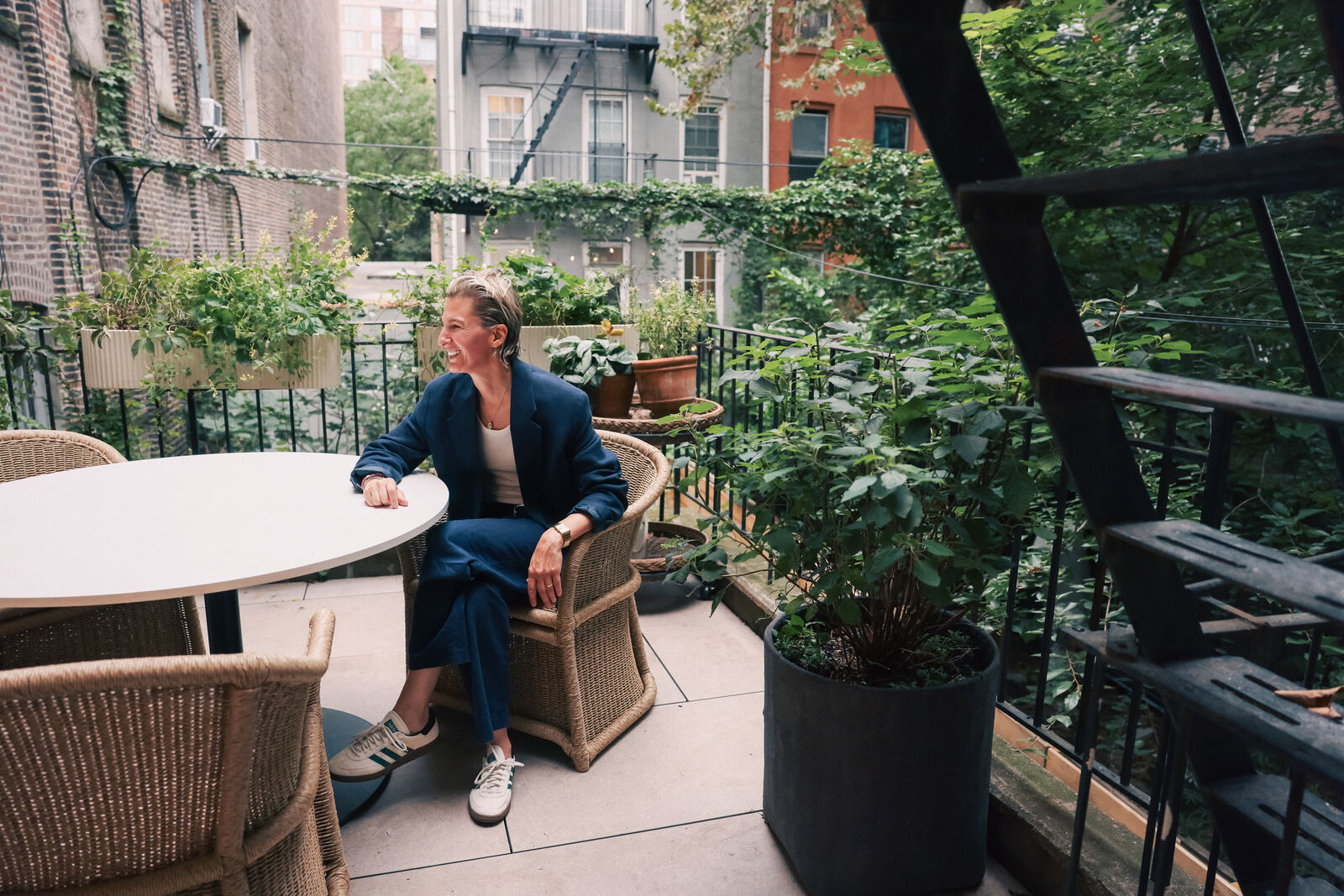 Albi Zhubi seated in a wicker chair at a round table on a leafy rooftop terrace, fire escapes behind her